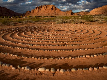 The image depicts a desert landscape with concentric circles made of stones, set against a backdrop of rocky mountains under a cloudy sky.