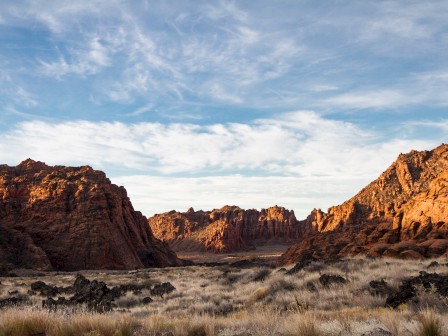 The image shows a desert landscape with rocky red cliffs, dry grass, and a partly cloudy sky at sunset or sunrise, casting warm light over the scene.