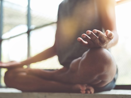 A person is meditating in a cross-legged position indoors, with one hand resting on their knee and sunlight streaming in through a window.