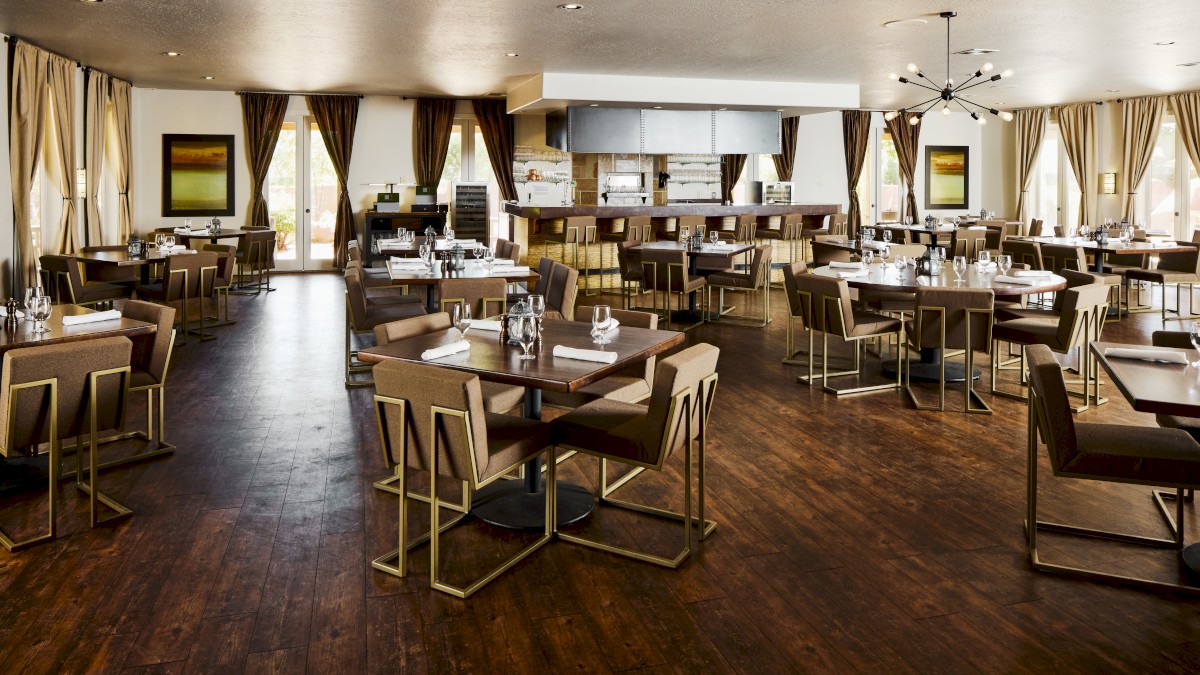 An elegant, modern restaurant with wooden floors, neatly arranged tables, and large windows draped with curtains.