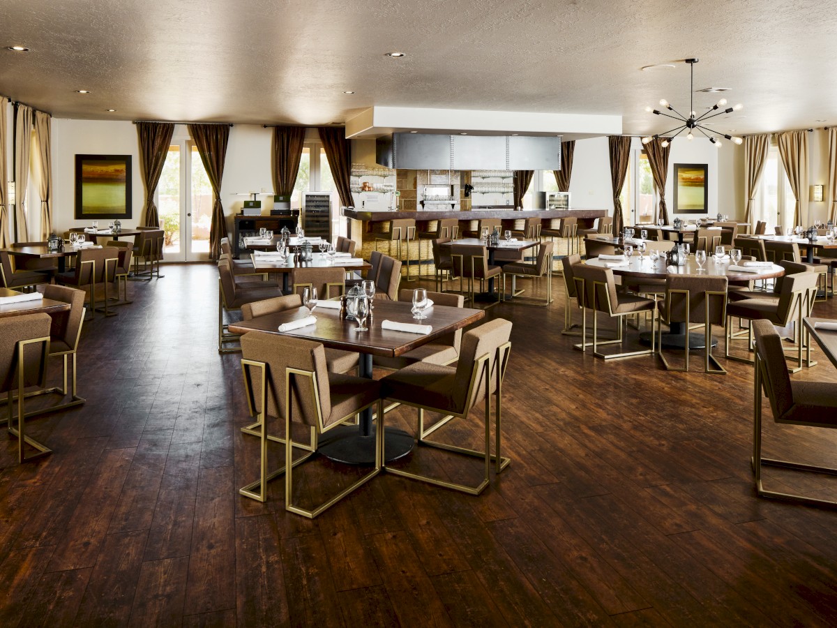 An elegant, modern restaurant with wooden floors, neatly arranged tables, and large windows draped with curtains.