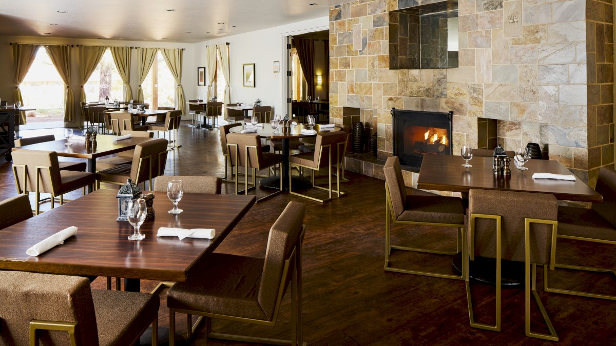 A cozy, modern restaurant with neatly arranged tables and chairs, a stone fireplace, and large windows with curtains. Elegant and inviting ambiance.