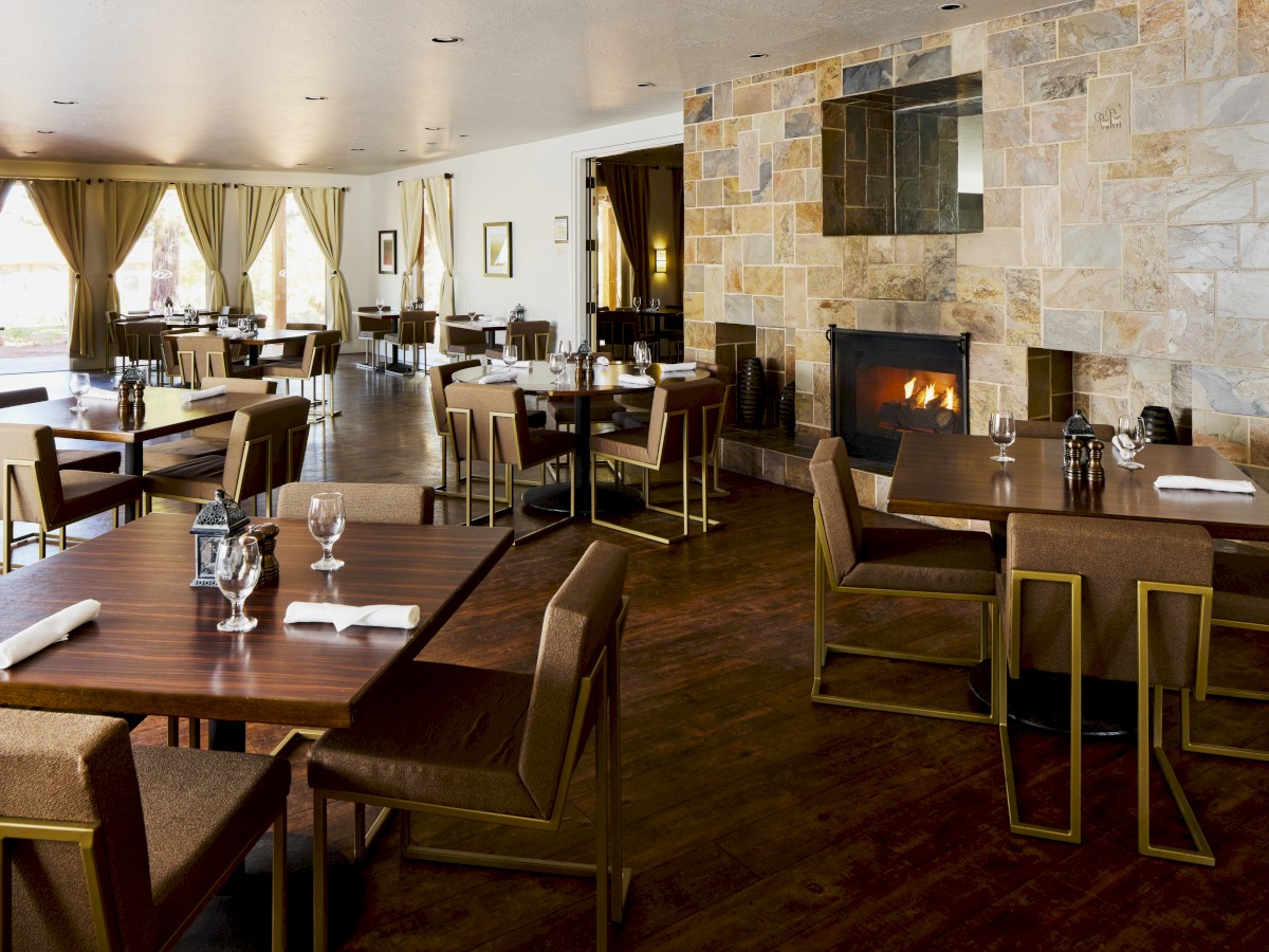 A cozy, modern restaurant with neatly arranged tables and chairs, a stone fireplace, and large windows with curtains. Elegant and inviting ambiance.