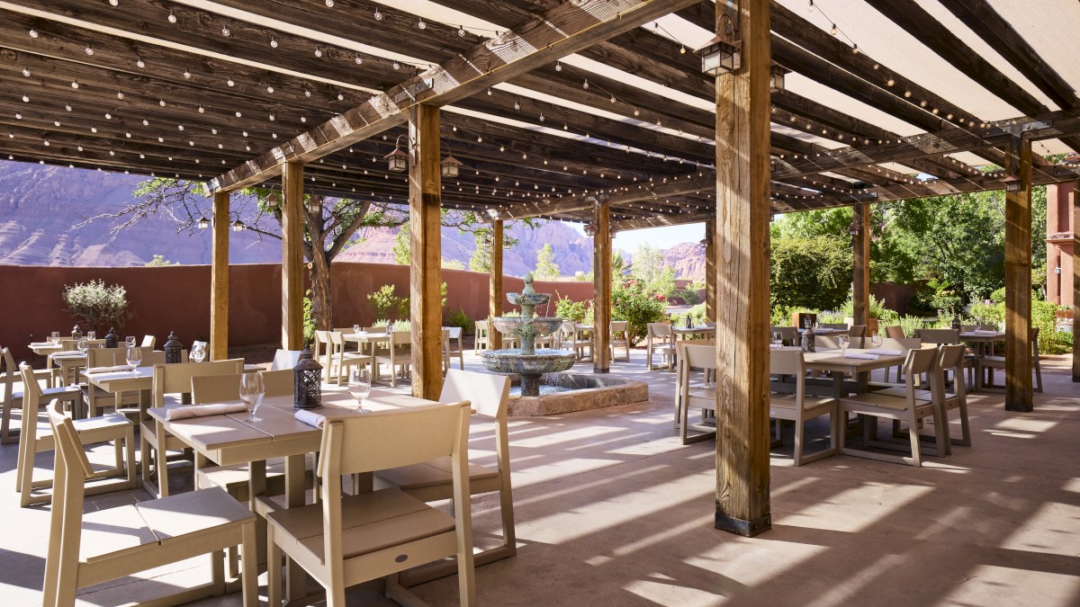 An outdoor patio with wooden pergolas, tables, and chairs is set for dining. Strings of lights hang above, and there is a fountain in the background.