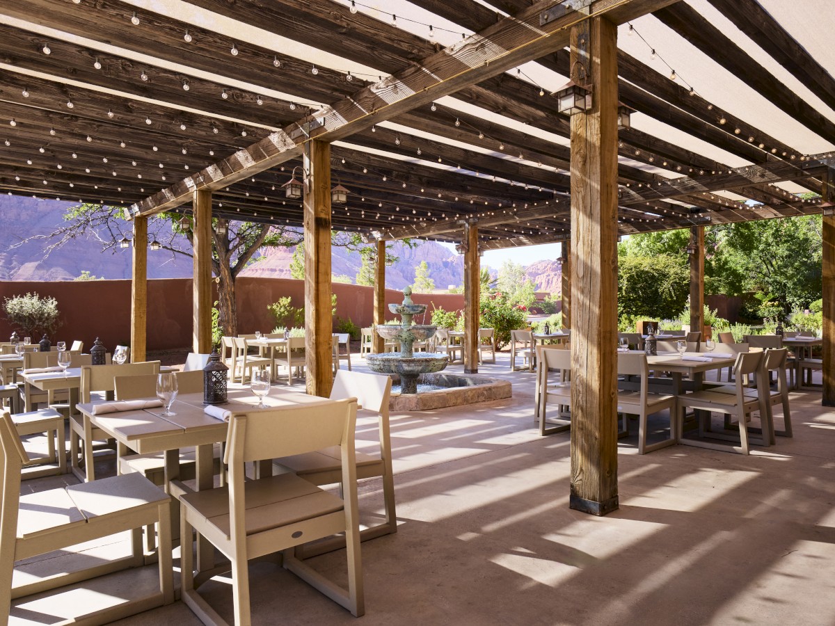 An outdoor patio with wooden pergolas, tables, and chairs is set for dining. Strings of lights hang above, and there is a fountain in the background.