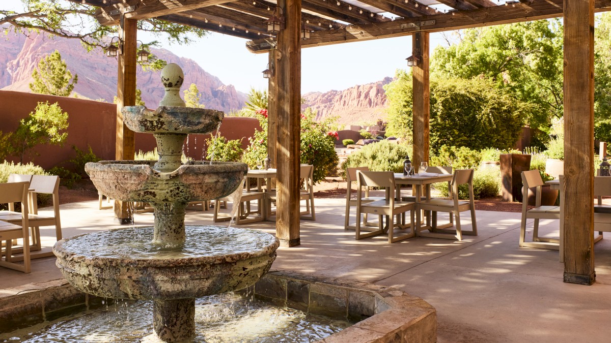 An outdoor patio with a fountain, wooden pergola, and dining tables overlooks a desert landscape and mountainous scenery, surrounded by lush greenery.