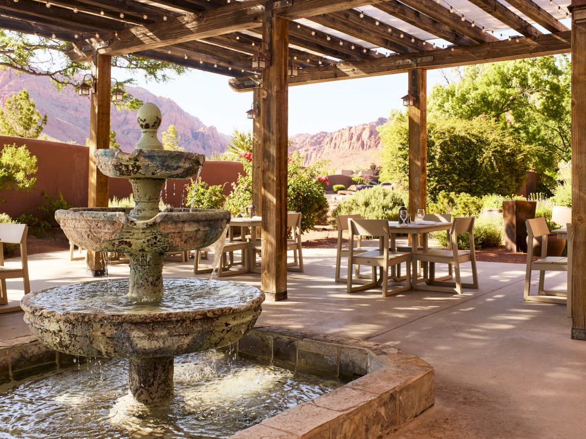 An outdoor patio with a fountain, wooden pergola, and dining tables overlooks a desert landscape and mountainous scenery, surrounded by lush greenery.