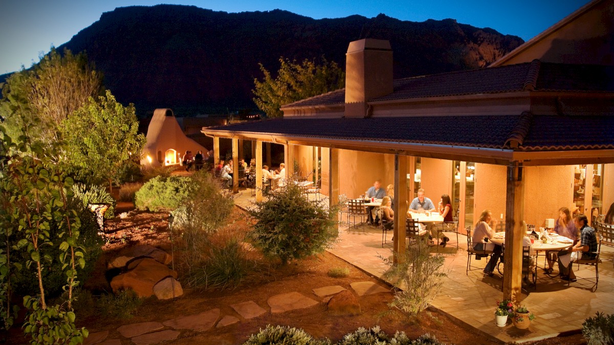 An outdoor restaurant at dusk, featuring tables with people dining on a covered patio surrounded by lush greenery and a mountainous backdrop.