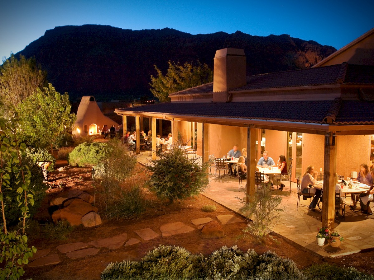 An outdoor restaurant at dusk, featuring tables with people dining on a covered patio surrounded by lush greenery and a mountainous backdrop.