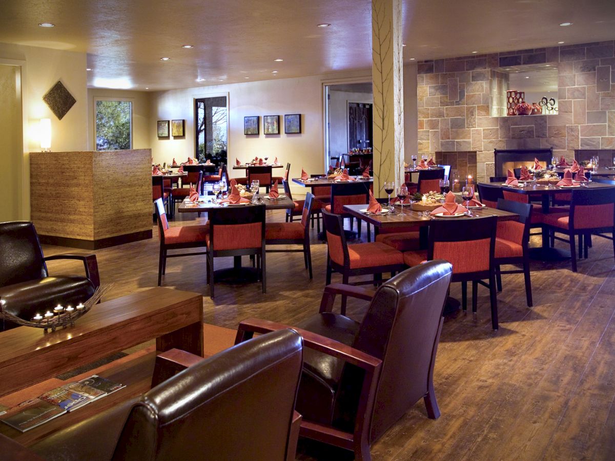 A cozy restaurant interior with wooden furniture, set tables, leather chairs, and a stone fireplace in the background, creating a warm atmosphere.