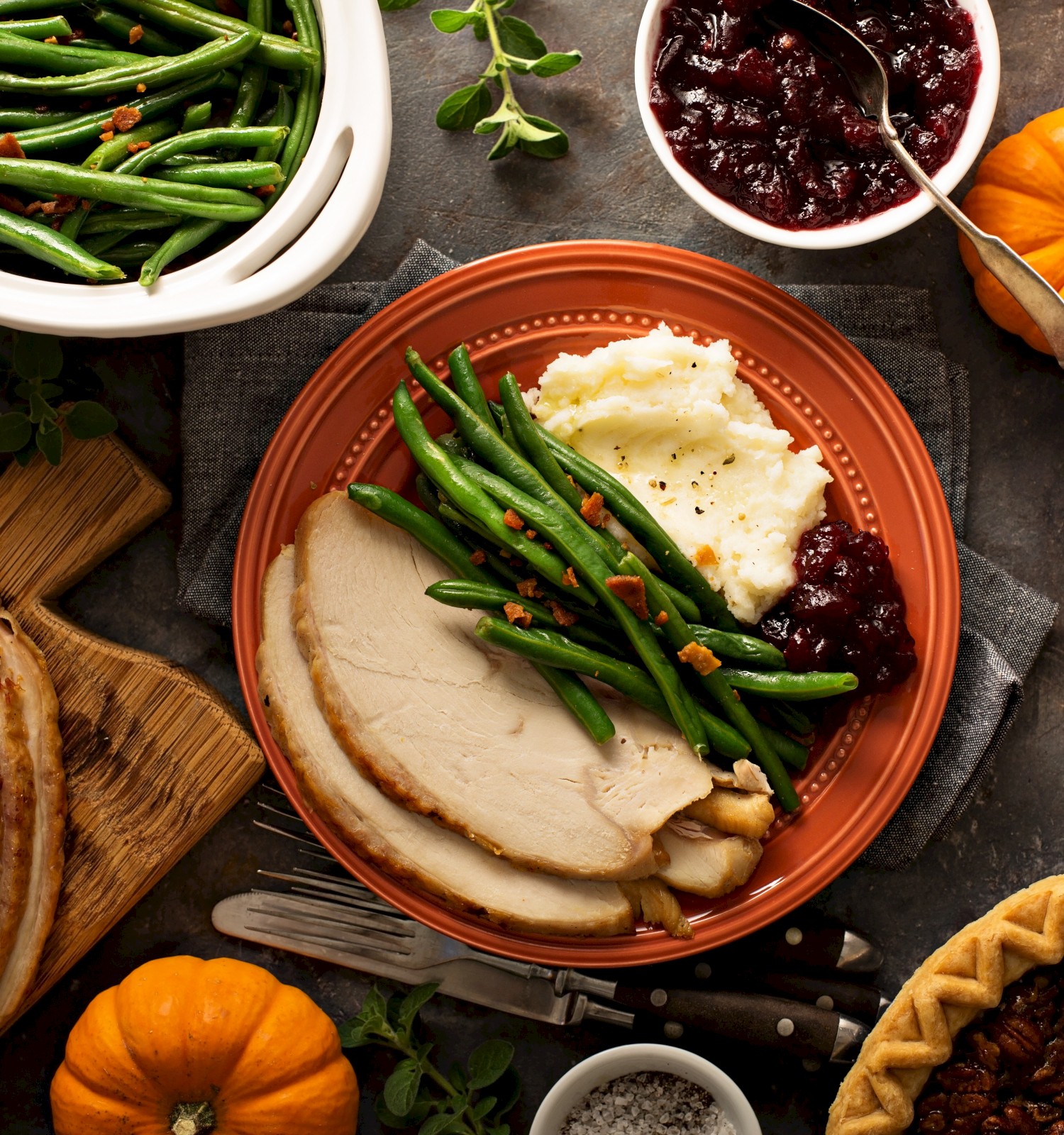 A Thanksgiving meal with turkey, green beans, mashed potatoes, cranberry sauce, a pecan pie, and small pumpkins on a table.