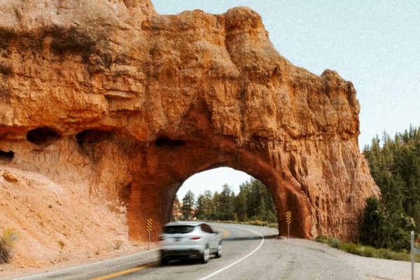 A car drives through a natural rock arch on a winding road with trees in the background under a clear sky.