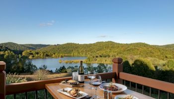 A table set with food and wine on a balcony overlooks a scenic lake and forested hills under a clear blue sky, creating a peaceful dining setting.