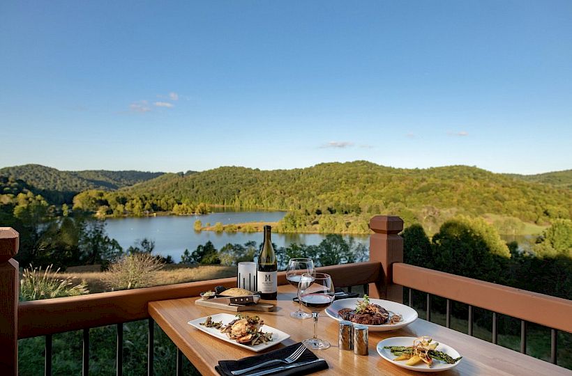 A table set with food and wine on a balcony overlooks a scenic lake and forested hills under a clear blue sky, creating a peaceful dining setting.