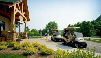 The image shows a man walking towards a building carrying a golf bag, while two people sit in golf carts on a paved area with landscaping.