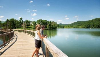 Two people stand on a wooden boardwalk, looking out over a serene lake with lush green hills and a clear blue sky in the background.