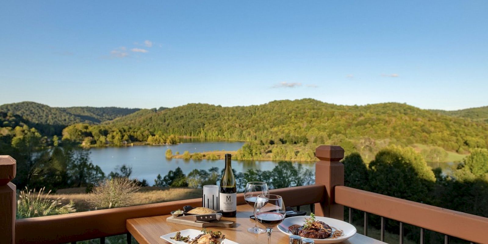 A table with three plates of food, a wine bottle, and glasses overlooks a scenic lake surrounded by trees and hills under a clear blue sky.