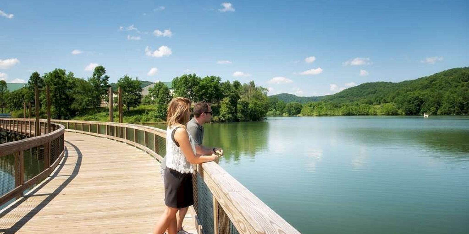 A couple stands on a wooden boardwalk overlooking a serene lake surrounded by trees and rolling hills under a clear blue sky with a few clouds.