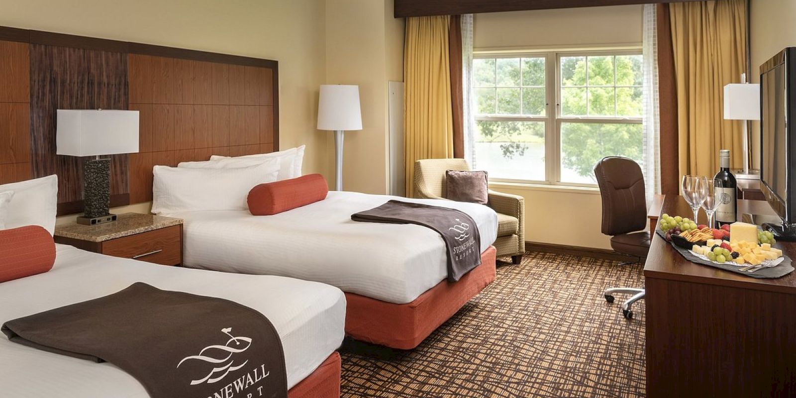 This image shows a hotel room with two double beds, a large window with curtains, a TV, and a table with fruit and wine.