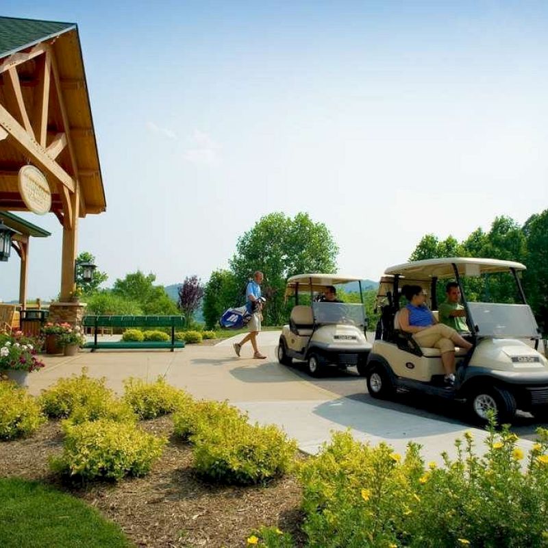 The image shows two people in golf carts near a building with wooden beams and landscaped greenery, while a person walks carrying golf clubs.