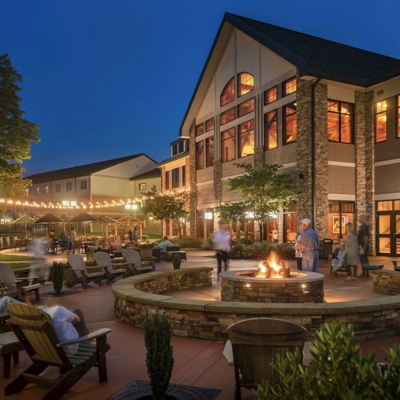 An evening scene of a resort with outdoor seating, people around a central fire pit, string lights overhead, and a large building in the background.