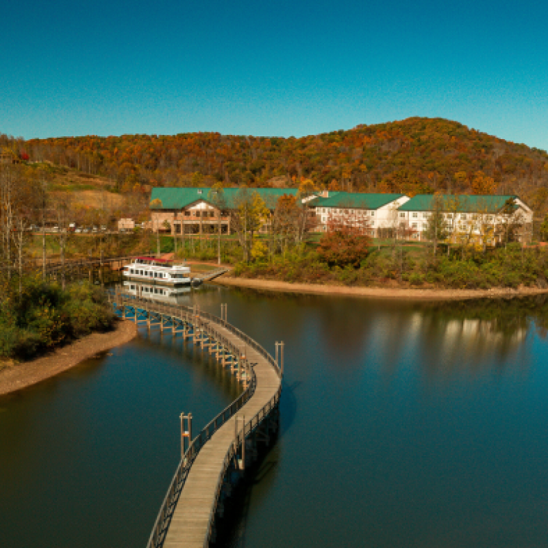 A serene scene of a lake with a boat dock, a curved walkway leading to buildings with green roofs, surrounded by autumn foliage and hills.