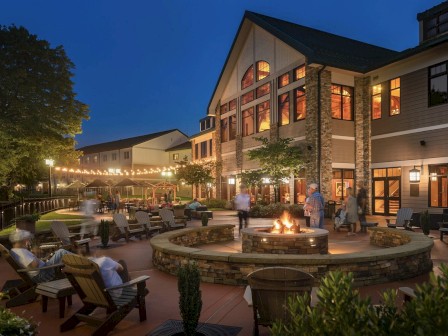 A cozy outdoor area with a fire pit, seating, string lights, and people enjoying the evening next to a large, illuminated building.