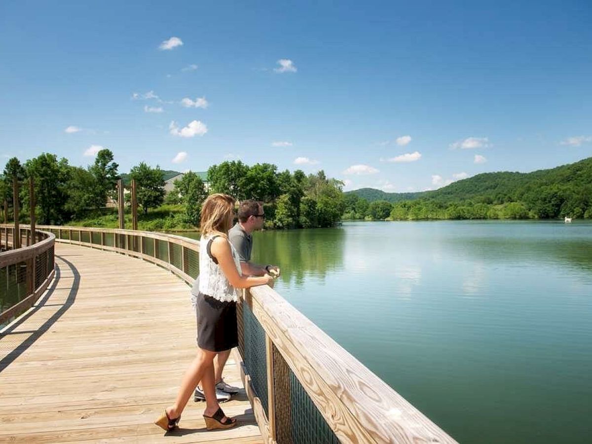 A couple stands on a wooden walkway overlooking a calm lake surrounded by greenery under a clear blue sky with a few scattered clouds.