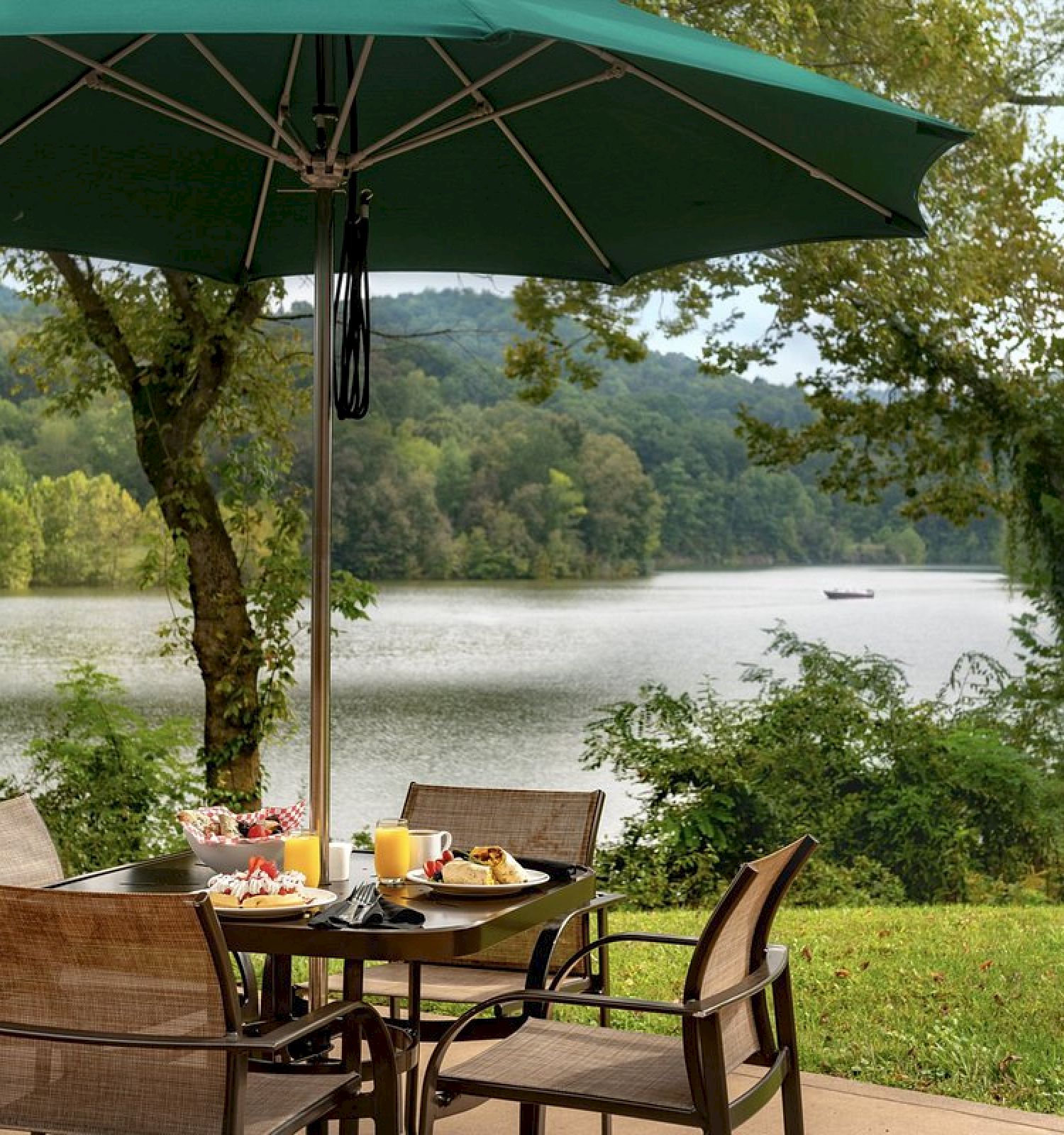 A patio dining set with a green umbrella, set for breakfast, overlooks a serene lake surrounded by lush trees and hills, creating a peaceful scene.