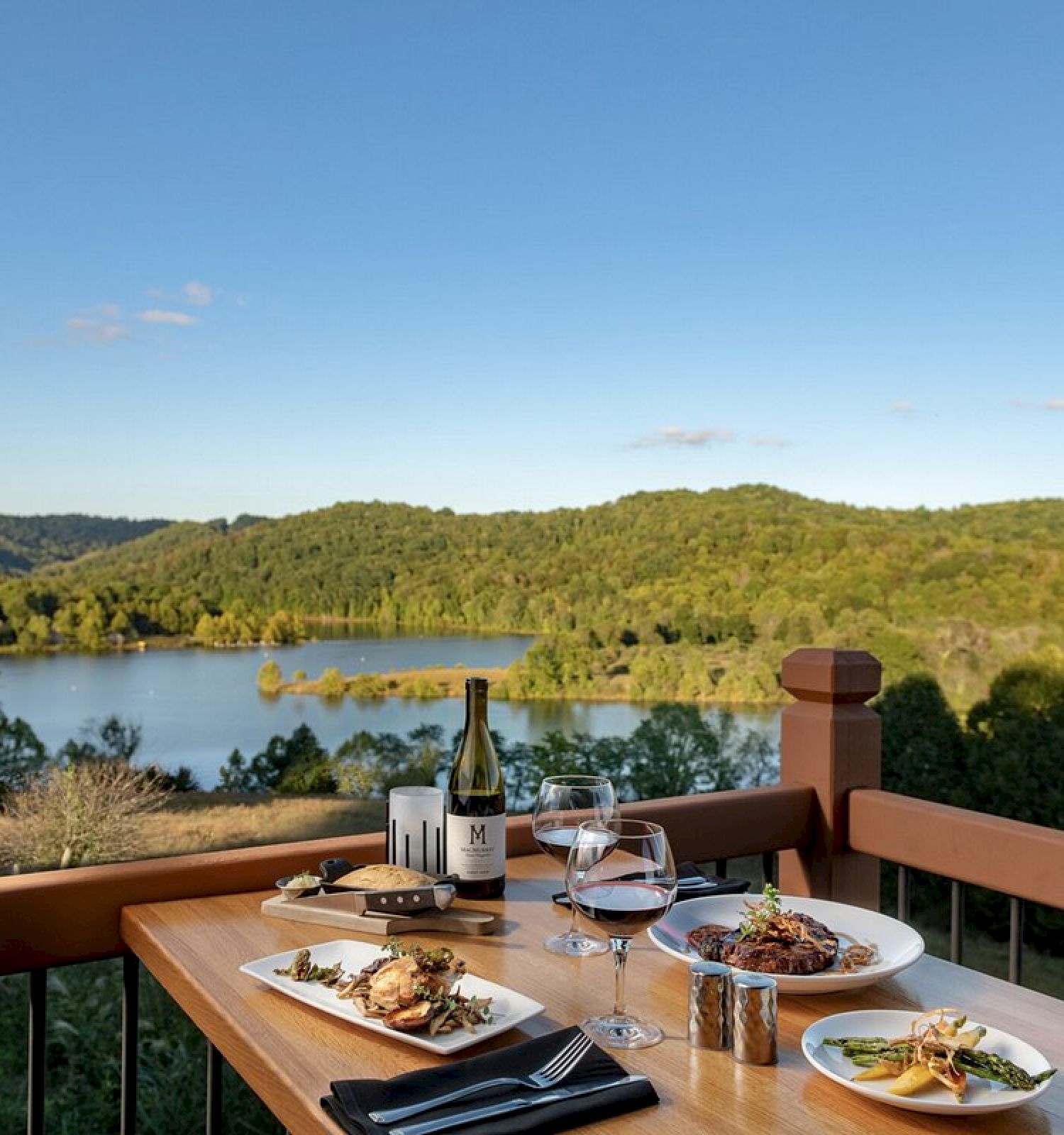 A scenic outdoor dining table with plated food, wine, and glasses set against a picturesque mountain and lake view in the background.