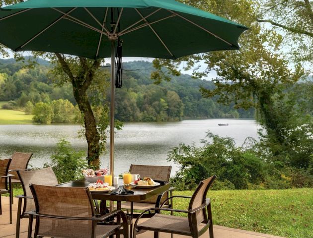 A scenic outdoor dining area with a table set for four under a green umbrella, adjacent to a lake with a picturesque view of trees and hills.