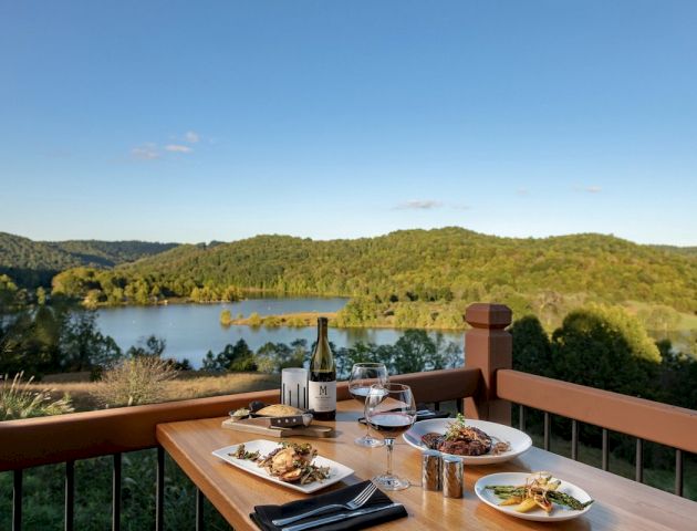 A table is set with food, wine, and glasses on a deck overlooking a scenic view of a lake and forested hills under a clear blue sky.