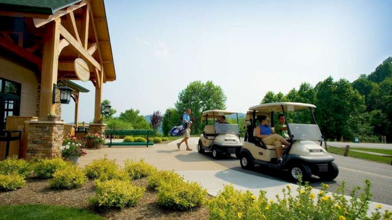 A person is walking towards a building, while two golf carts with passengers are on a path nearby, surrounded by greenery and flowers.