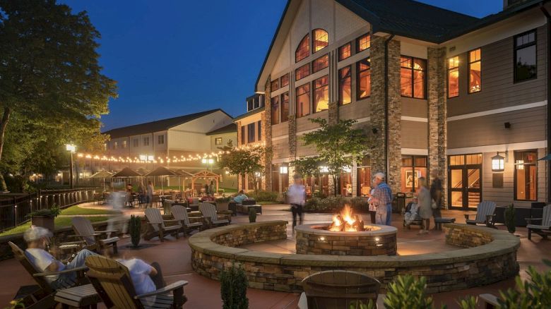 The image shows an outdoor area at night with people sitting around a large fire pit. The area is decorated with string lights and surrounded by buildings.